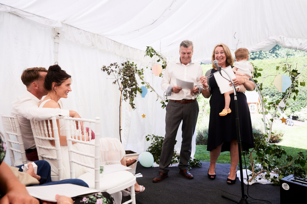 Happy grandparents hold a baby at the front of the ceremony room, doing a reading and smiling