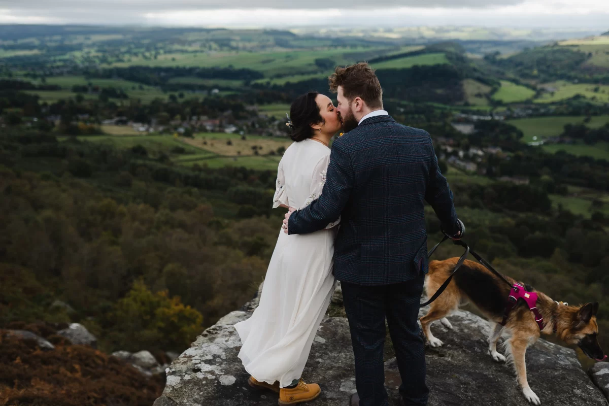A bride and groom kiss, standing on an outcrop high above a green landscape with their dog