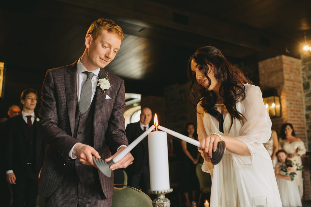 A bride and groom light a large candle from two long tapered candles. They look serious as they bring the flames together.