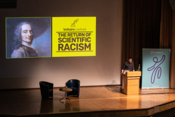 Adam Rutherford begins his Voltaire Lecture, whose title is displayed behind him alongside an illustration of Voltaire