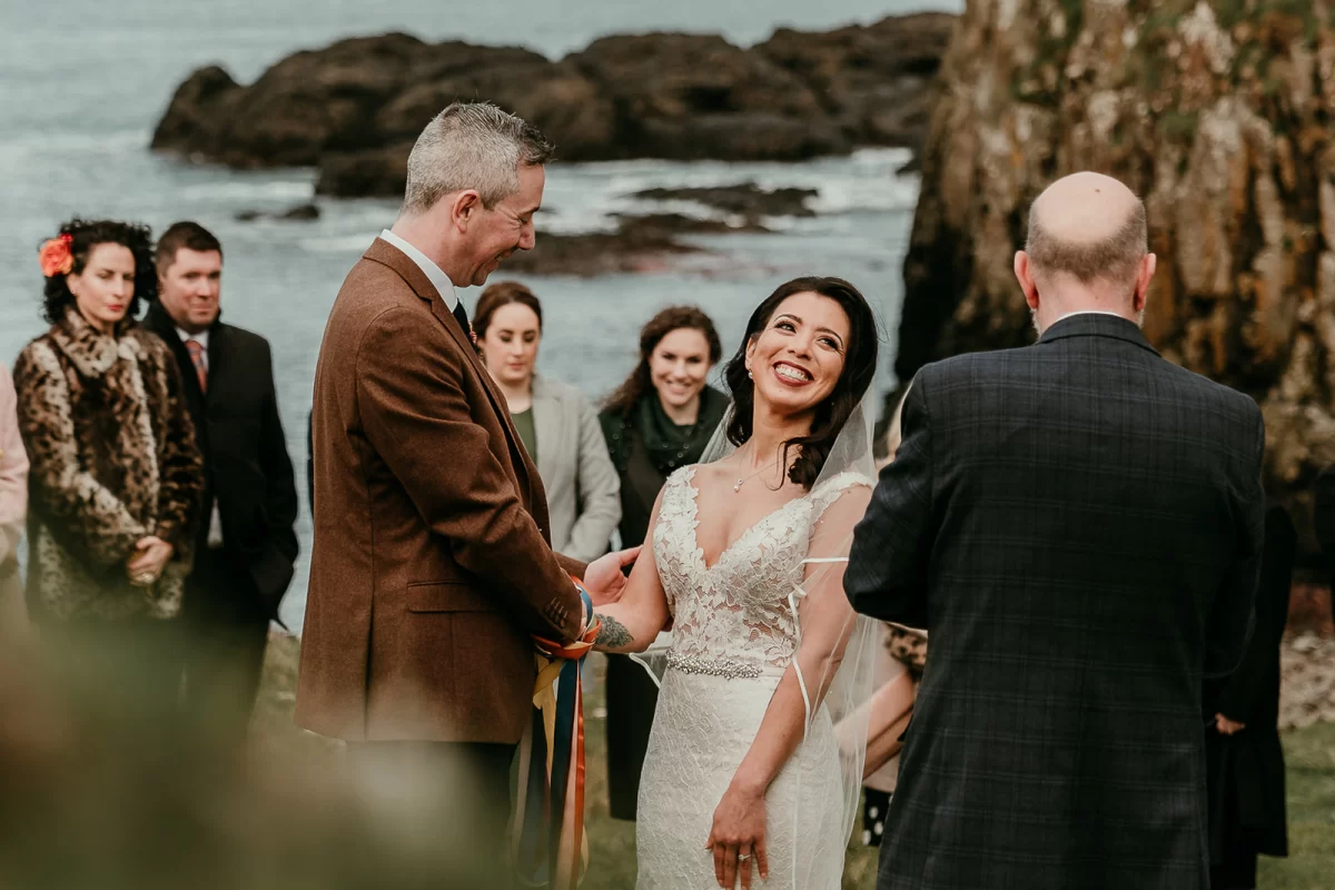 A moment from the ceremony, the bride grinning at the celebrant as she holds her groom's hand. The wedding guests behind wrapped up warm with the sea and cliffs behind them.
