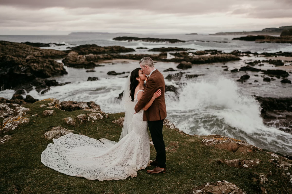 Bride and groom together, locked in an embrace. They stand in front of a wild coastline with the sea crashing on the rocks behind them.