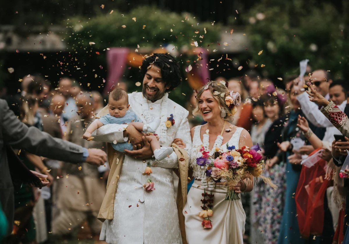 Lucia and Amit walk down the aisle after their ceremony, holding baby Arti, surrounded by confetti and smiling with joy