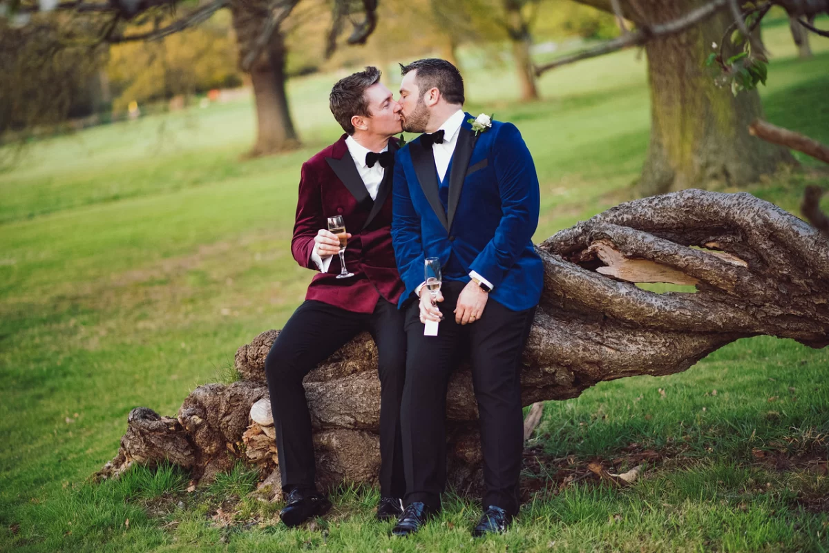 Two men in velvet tuxedo jackets kiss outside on their big day