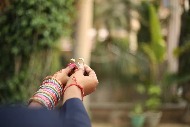 Two hands holding rings, trees in the background. The bride's wrist is full of colourful bracelets and we can see henna on her hand.