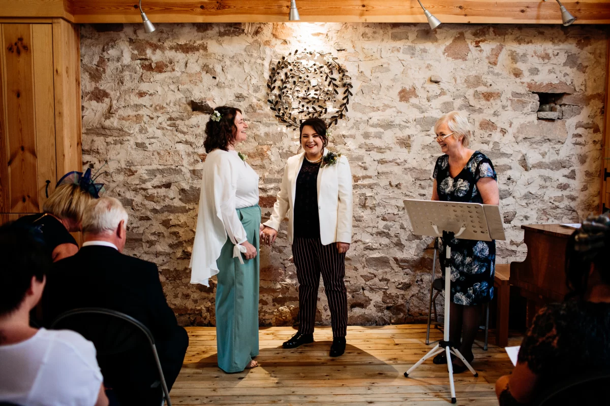 A wedding ceremony with two brides, one wearing loose flowing sage green trousers and a floaty white top and the other a smart cream blazer and pinstriped trousers. They are smiling with the celebrant.