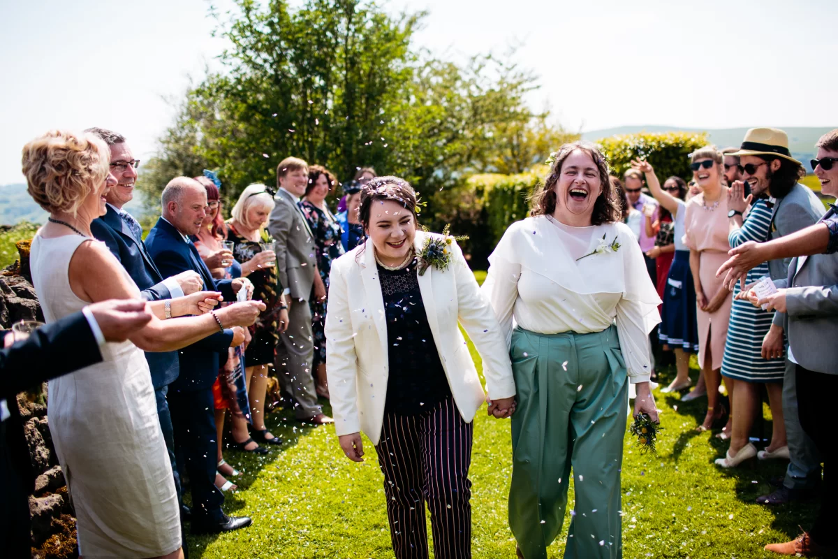 Two brides walk down a green path, with friends and family lining the way and throwing confetti. It is joyous and full of sunshine.