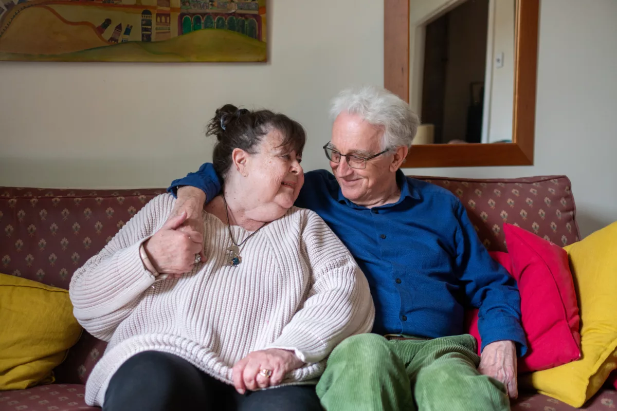 An older couple sit together on the sofa, cosy in jumpers and smiling at each other
