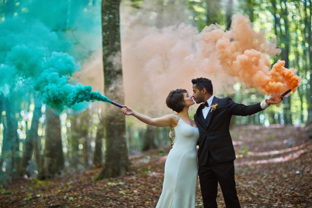 A bride and groom hold colourful smoke in each hand as they kiss in the woods