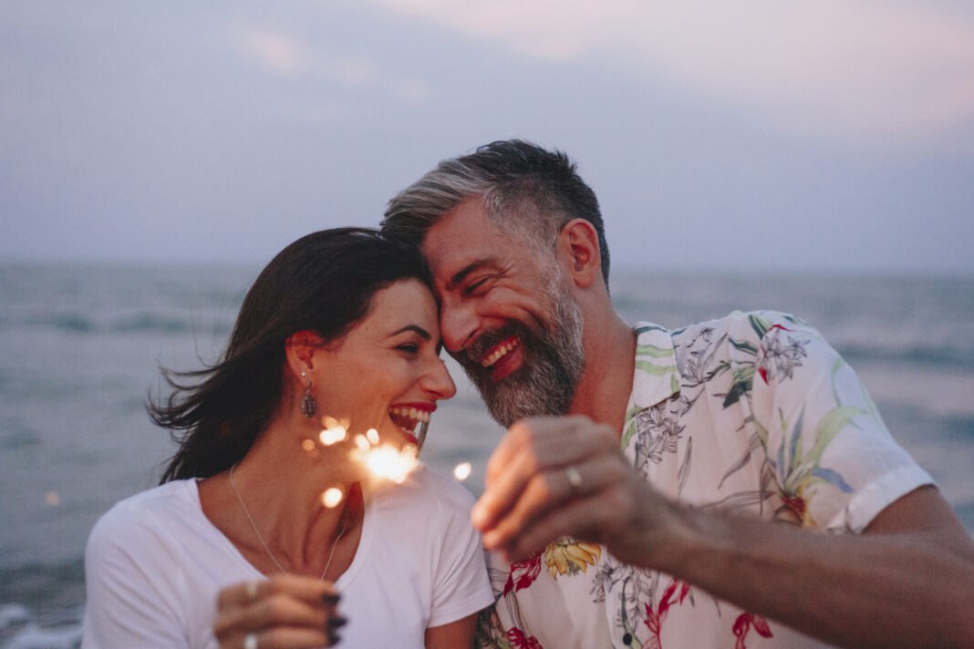 A man and woman on a beach, the sea behind them, lighting sparklers and sharing a moment together full of joy and love