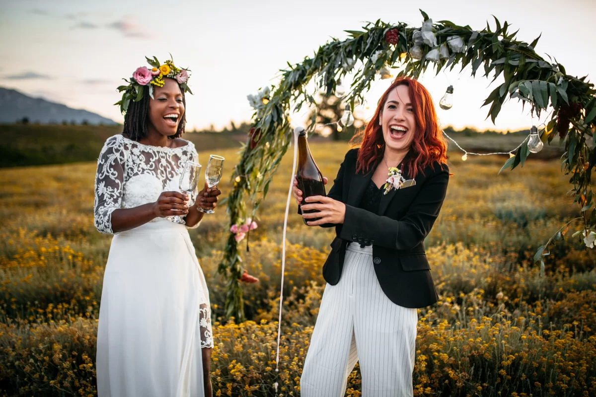 Two brides celebrate outside, opening a bottle of champagne and laughing. 