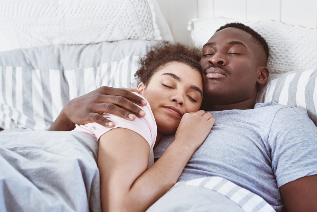A young Black couple lie cosily in bed, snuggled together wearing t-shirts