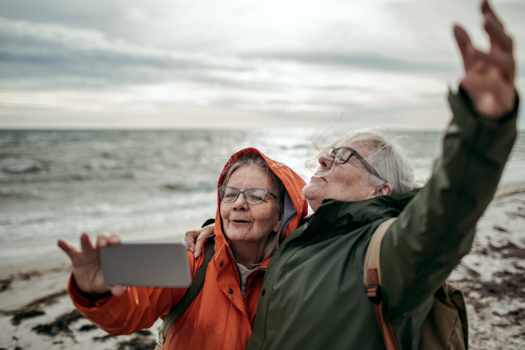 An older lesbian couple in raincoats on the beach, arms outstretched and head thrown back for a selfie