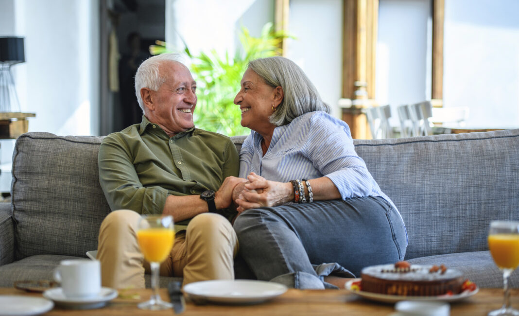 An older couple on the sofa, grinning mischievously at each other and holding hands
