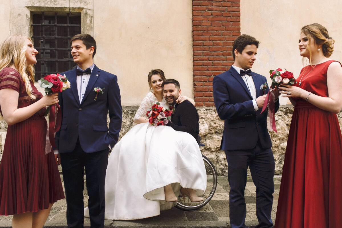 A young newly married couple, surrounded by their grooms and bridesmaids. The groom is a wheelchair user and the bride is sitting on his lap with her arms around his neck.